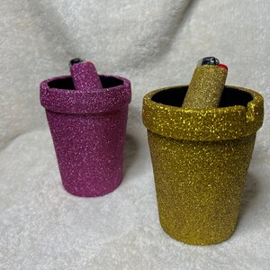 May include: Two glitter-covered ceramic ashtrays, one pink and one gold, with lighters inside.