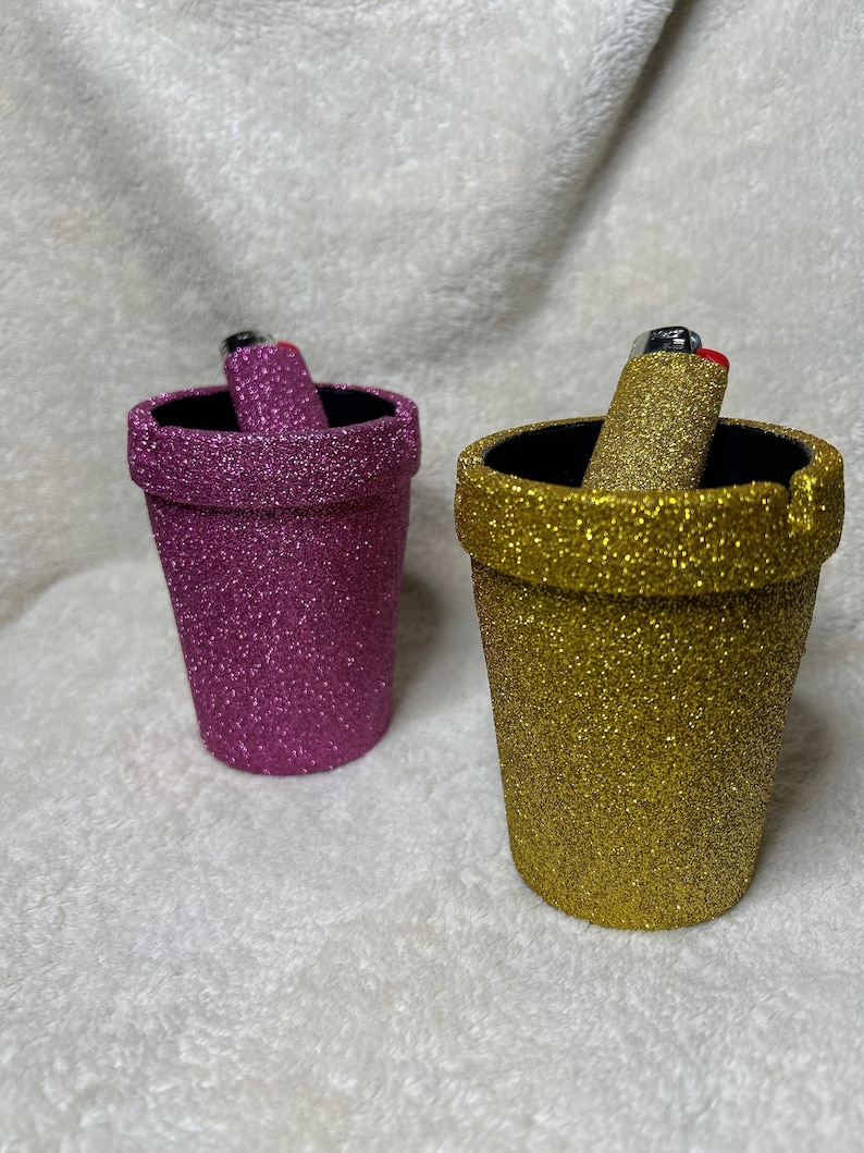 May include: Two glitter-covered ceramic ashtrays, one pink and one gold, with lighters inside.