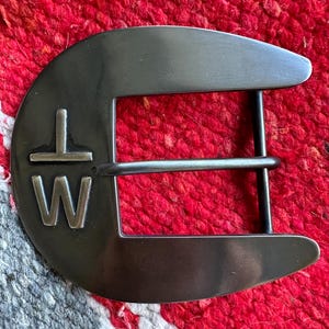 May include: A silver metal belt buckle with the letters "T W" engraved on the front. The buckle is a traditional style with a rounded top and a single prong.