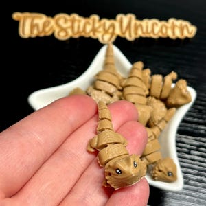 May include: A collection of small, tan-colored, segmented lizard figurines. One is held in a hand, showcasing its detailed design with small eyes. The figurines are in a white star-shaped dish, with the text "The Sticky Unicorn" in the background.