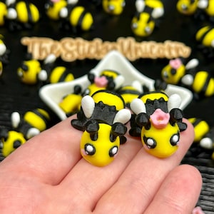 May include: A collection of yellow and black bee-shaped figurines, some with pink flower accents, are displayed. The figurines are held in a hand, with more bees scattered around. The text "The Sticker Unicorn" is visible in the background.
