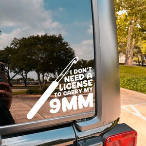 May include: White vinyl decal on a vehicle window. The decal features a crochet hook and the text: "I DON'T NEED A LICENSE TO CARRY MY 9MM."