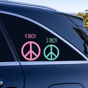 May include: Two peace sign decals on a black car window. The larger decal is 4 inches and pink. The smaller decal is 3 inches and green.