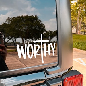 May include: White "WORTHY" decal with a cross design, applied to a vehicle window. The decal is in a handwritten style, with the word "WORTHY" in bold letters. The background shows trees, sky, and a glimpse of a person holding a phone.