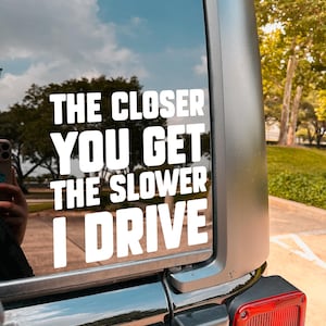 May include: A white vinyl decal on a vehicle's rear window. The text reads "THE CLOSER YOU GET THE SLOWER I DRIVE" in a bold, sans-serif font. The background shows a blue sky with clouds and trees.