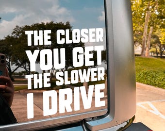 May include: A white vinyl decal on a vehicle's rear window. The text reads "THE CLOSER YOU GET THE SLOWER I DRIVE" in a bold, sans-serif font. The background shows a blue sky with clouds and trees.