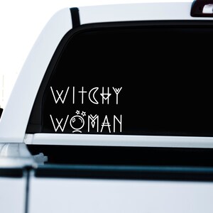 May include: A white truck with a black rear window. The window has a white vinyl decal that reads "Witchy Woman" with a crescent moon and a crystal ball graphic.
