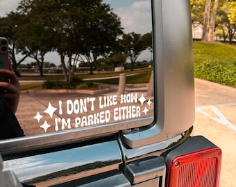 May include: White vinyl decal on a vehicle window. The decal reads "I DON'T LIKE HOW I'M PARKED EITHER" in a playful font, with starburst accents. The image shows a partial view of the vehicle's exterior, with a reflection of trees and sky.