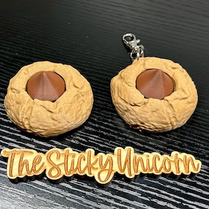 May include: Two tan-colored, round keychains with a dark brown, cone-shaped center. One keychain has a silver clasp. The keychains are next to a wooden sign that reads "The Sticky Unicorn".
