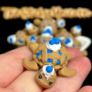 May include: A miniature, tan turtle figurine with blue and white accents. The turtle's shell has blue spots, and it has a white and blue topping on its head and face. The figurine is held in a hand against a black background.