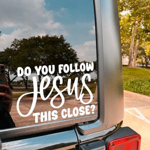 May include: White vinyl decal on a vehicle window. The decal reads "DO YOU FOLLOW JESUS THIS CLOSE?" in a mix of block and cursive fonts. The background shows a blue sky with clouds and trees.