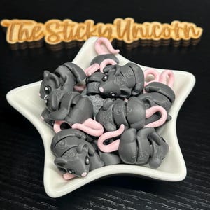 May include: A white star-shaped dish filled with gray mouse-shaped figurines with pink tails. The figurines have small black eyes and are arranged in a pile. The wooden sign above the dish reads "The Sticky Unicorn".