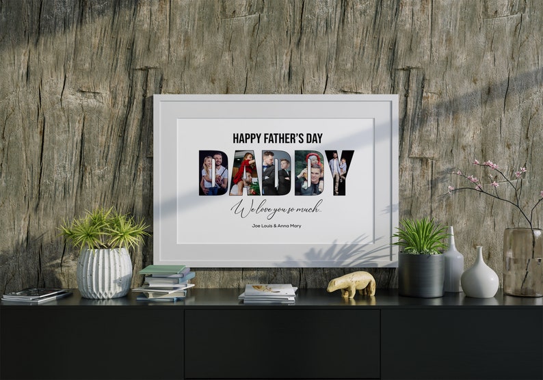 May include: A framed print with the text "HAPPY FATHER'S DAY DADDY We love you so much" and family photos inside the word "DADDY". The print is displayed on a black cabinet with decorative items and plants.