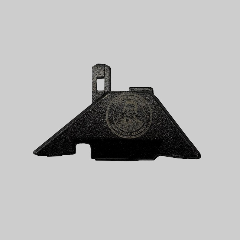 Puede incluir: Un soporte met&aacute;lico negro texturizado con una forma &uacute;nica. Presenta una secci&oacute;n elevada con una abertura cuadrada y un emblema circular con texto y un retrato. El texto "FAYETTEVILLE, ARKANSAS" es visible.