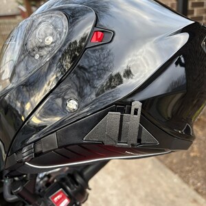 Puede incluir: Casco de motocicleta negro brillante con una visera transparente. El casco tiene un bot&oacute;n rojo y un ribete negro. El casco est&aacute; montado en una motocicleta roja.