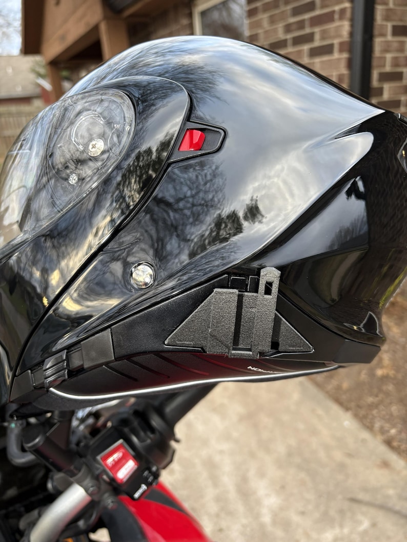 Puede incluir: Casco de motocicleta negro brillante con una visera transparente. El casco tiene un bot&oacute;n rojo y un ribete negro. El casco est&aacute; montado en una motocicleta roja.