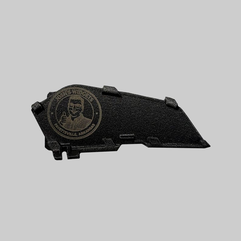 May include: Black, textured, angular widget with the text "Josh's Widgets Fayetteville, Arkansas" in a circular emblem. The emblem features a man giving a thumbs up. The widget has several raised rectangular features.
