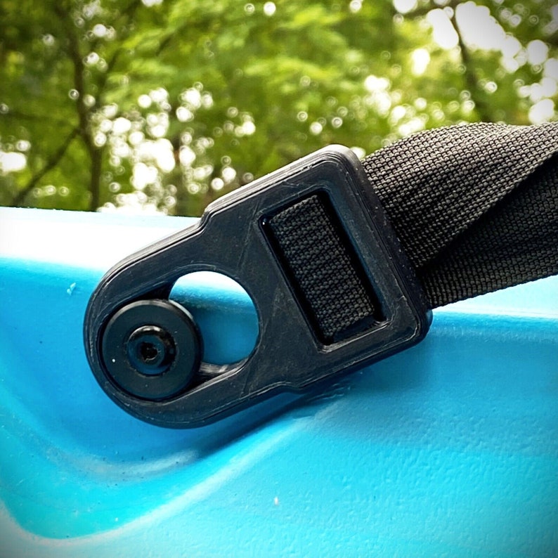 Op de afbeelding: Close-up van een zwarte plastic gesp met een metalen ring. De gesp is bevestigd aan een zwarte nylon band. De gesp bevindt zich op een blauw oppervlak.