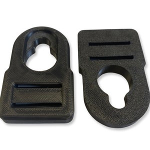 Op de afbeelding: Twee zwarte plastic haken met een rechthoekige vorm en een rond gat in het midden. Elke haak heeft twee horizontale gleuven aan de bovenkant.