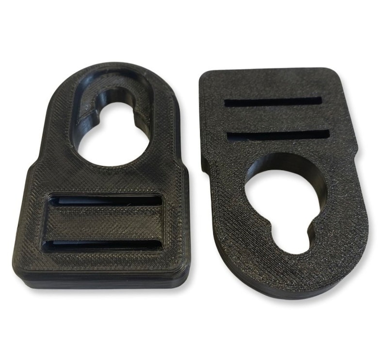 Op de afbeelding: Twee zwarte plastic haken met een rechthoekige vorm en een rond gat in het midden. Elke haak heeft twee horizontale gleuven aan de bovenkant.