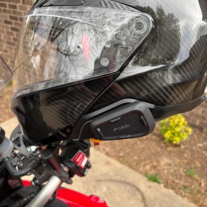 May include: A black motorcycle helmet with a clear visor and a carbon fibre weave pattern. A black Cardo communication device is attached to the side of the helmet. The helmet is on a red motorcycle.