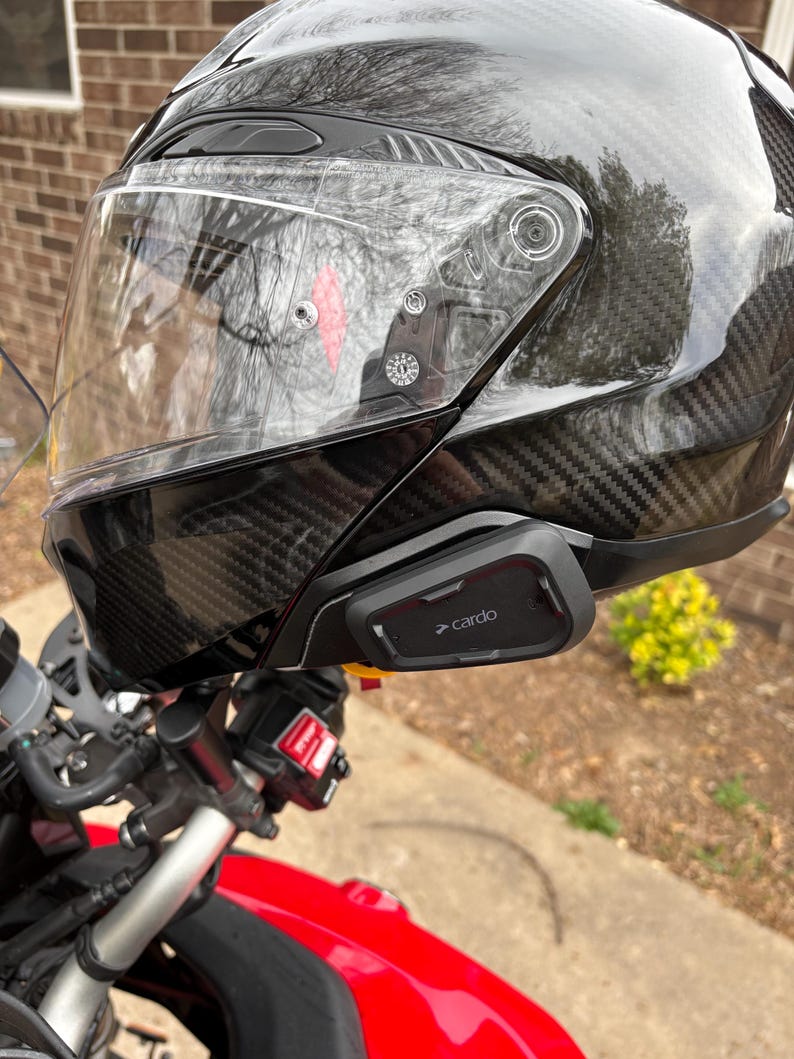 May include: A black motorcycle helmet with a clear visor and a carbon fibre weave pattern. A black Cardo communication device is attached to the side of the helmet. The helmet is on a red motorcycle.