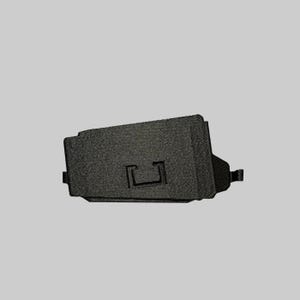 May include: A black, textured, rectangular automotive brake pad component featuring a central U-shaped indentation on its flat surface. The object has a rigid, metallic appearance with small protruding tabs on the left and right sides.