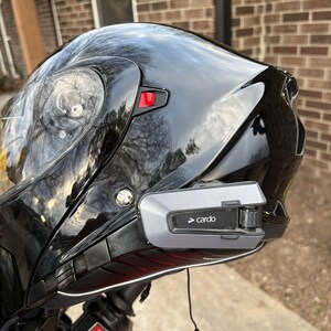 Puede incluir: Casco de moto negro brillante con visera transparente y sistema de comunicaci&oacute;n Cardo adjunto. El dispositivo Cardo es gris y negro con el nombre de la marca impreso. El casco est&aacute; montado en una moto roja.