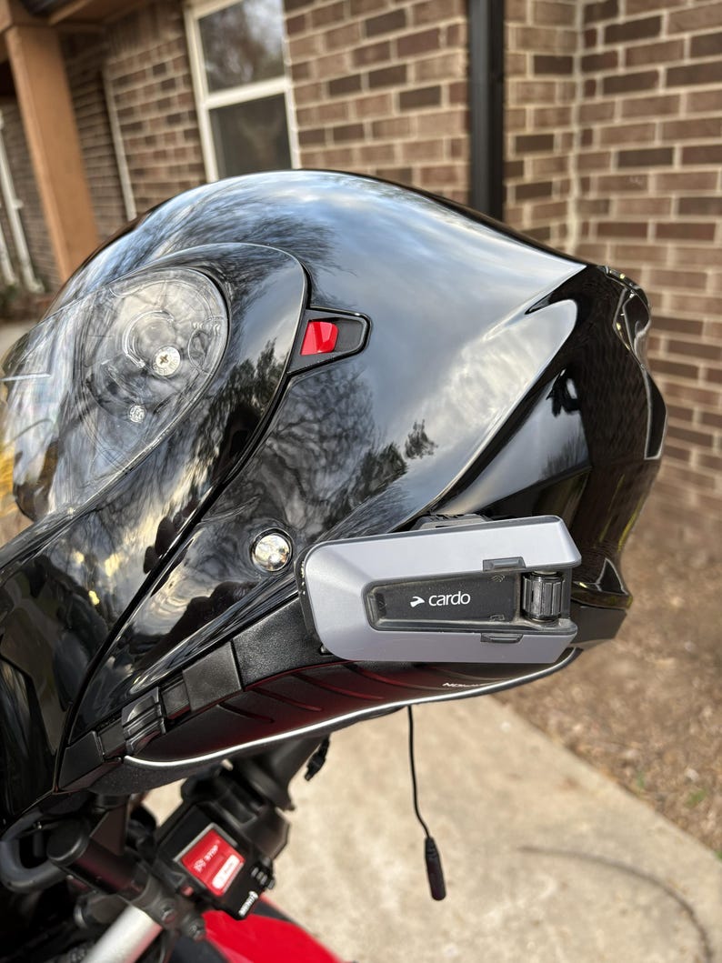 Puede incluir: Casco de moto negro brillante con visera transparente y sistema de comunicaci&oacute;n Cardo adjunto. El dispositivo Cardo es gris y negro con el nombre de la marca impreso. El casco est&aacute; montado en una moto roja.