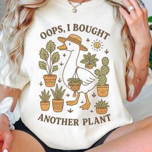 Puede incluir: Camiseta color crema con una ilustraci&oacute;n de un ganso con sombrero y plantas en macetas. El texto "OOPS, I BOUGHT ANOTHER PLANT" est&aacute; impreso encima y debajo del ganso. El dise&ntilde;o incluye un sol y varias plantas en macetas.