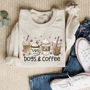 May include: A beige sweatshirt with a graphic of four coffee cups and paw prints. The text "Dog Mom" and "Dogs & Coffee" are printed on the sweatshirt.