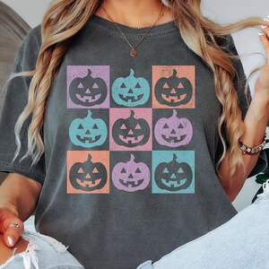 May include: Dark gray t-shirt featuring a grid of nine colorful jack-o'-lanterns. Each pumpkin is in a different color square: purple, blue, orange, and teal. The shirt is a comfortable, casual style.