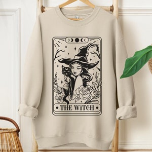 May include: A beige sweatshirt featuring a black and white illustration of a witch holding a black cat. The illustration is in the style of a tarot card and reads "THE WITCH" at the bottom.
