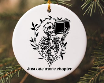 May include: White ceramic ornament with a black illustration of a skeleton reading a book, surrounded by leaves. The text "Just one more chapter" is printed below the illustration. A gold string is attached for hanging.