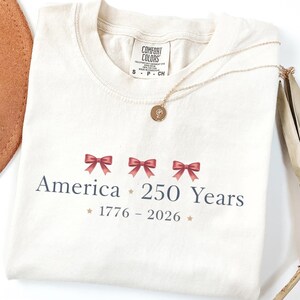 May include: Cream-colored t-shirt with the text "America 250 Years" and the years "1776-2026" printed in dark blue. The design includes three red bows. A gold necklace with a pendant is draped across the shirt.