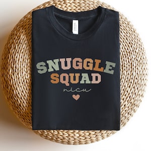 K&ouml;nnte beinhalten: Schwarzes T-Shirt mit dem Text "SNUGGLE SQUAD" in Regenbogenfarben und dem Wort "nicu" mit einem kleinen Herz darunter.