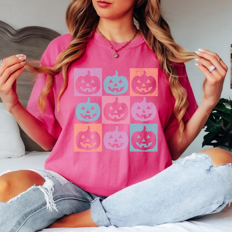 May include: A bright pink t-shirt featuring a grid of nine colorful jack-o'-lanterns. The pumpkins are in shades of pink, blue, orange, and purple. The shirt is a casual, comfortable style.