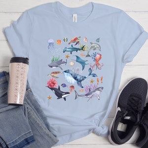 May include: Light blue t-shirt with a watercolor design of various sea creatures, including a whale, shark, octopus, jellyfish, and starfish.