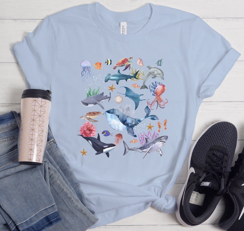 May include: Light blue t-shirt with a watercolor design of various sea creatures, including a whale, shark, octopus, jellyfish, and starfish.