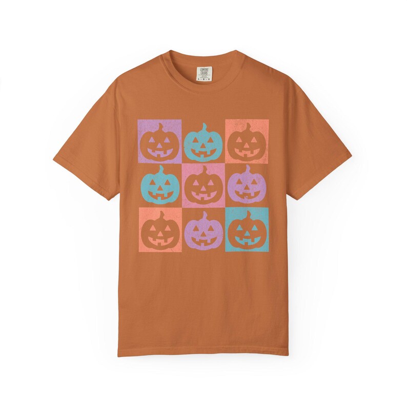 May include: A burnt orange t-shirt featuring a grid of nine colorful jack-o'-lanterns. Each pumpkin is set in a square of orange, purple, pink, and teal. The shirt is a short-sleeved crew neck.