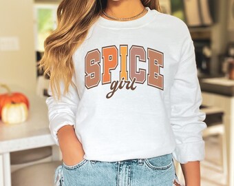 May include: A white crewneck sweatshirt featuring the text SPICE in a large, blocky, multi-colored varsity font with brown, orange, and tan letters, and the word girl written below in a brown cursive script font across the chest area.