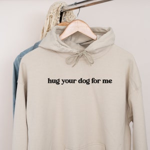 Pu&ograve; includere: Una felpa con cappuccio beige con una stampa nera che dice "hug your dog for me".