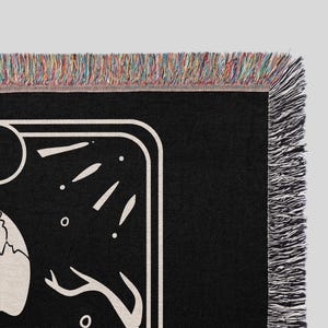 Puede incluir: Manta de tejido negro y blanco con un dise&ntilde;o de calavera y luna. La manta tiene un borde de flecos.