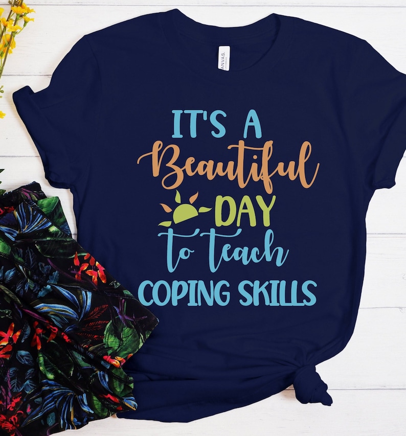 Pu&ograve; includere: T-shirt blu navy con la scritta "It's a Beautiful Day to Teach Coping Skills" in arancione, verde e blu. Il design include un sole. La maglietta &egrave; realizzata in materiale morbido.