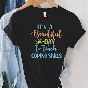 Pu&ograve; includere: T-shirt nera con la scritta "It's a Beautiful Day to Teach Coping Skills" in verde acqua, arancione e giallo. Il disegno include una grafica solare. La maglietta &egrave; appesa a un appendiabiti di legno.