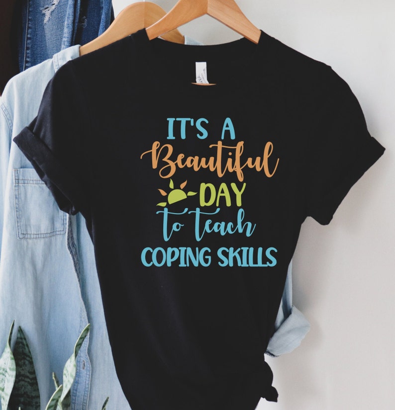 Pu&ograve; includere: T-shirt nera con la scritta "It's a Beautiful Day to Teach Coping Skills" in verde acqua, arancione e giallo. Il disegno include una grafica solare. La maglietta &egrave; appesa a un appendiabiti di legno.