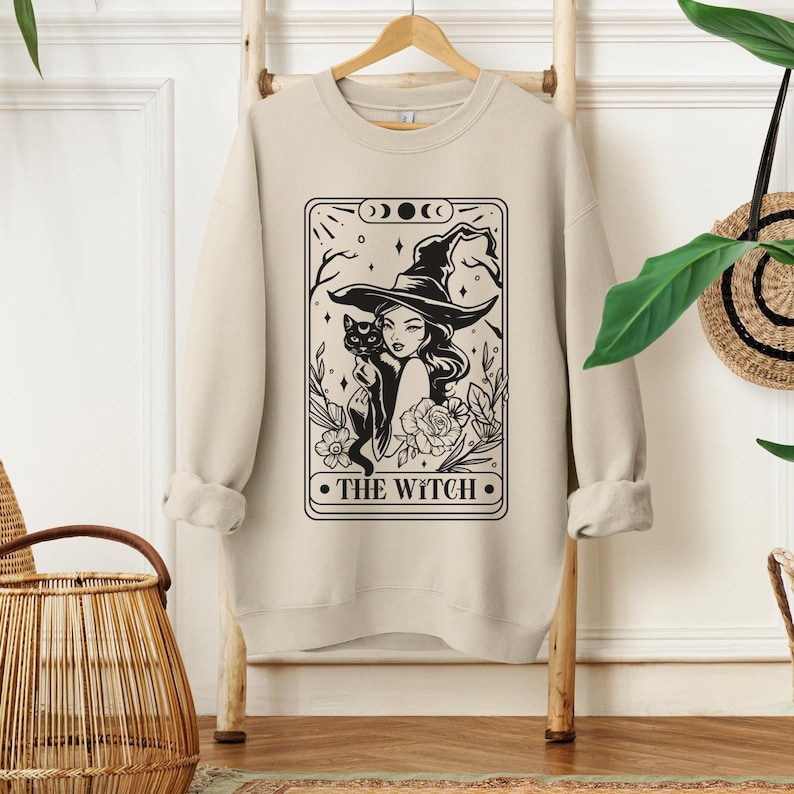 May include: A beige sweatshirt featuring a black and white illustration of a witch holding a black cat. The illustration is in the style of a tarot card and reads "THE WITCH" at the bottom.