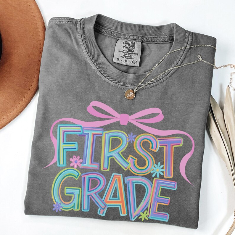 May include: A grey Comfort Colors t-shirt featuring the words "FIRST GRADE" in colourful, outlined letters. The design includes a pink bow and small flowers. A gold necklace with a pendant is draped across the shirt.