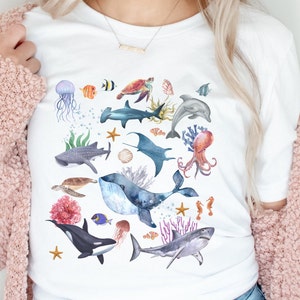 May include: White t-shirt with a colorful watercolor design of various sea creatures, including a whale, shark, octopus, seahorse, and starfish.