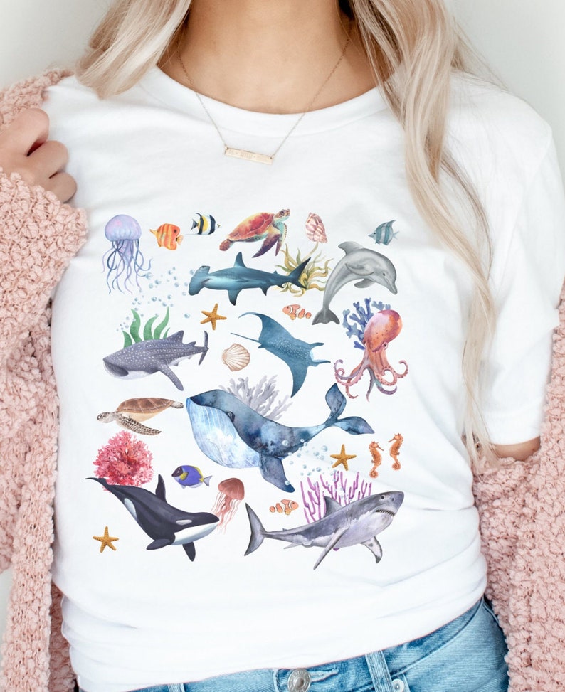 May include: White t-shirt with a colorful watercolor design of various sea creatures, including a whale, shark, octopus, seahorse, and starfish.